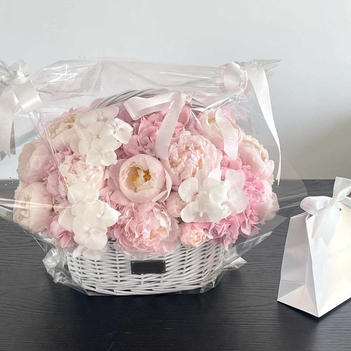 Pink and white preserved flower basket shown in clear wrapping beside a gift bag