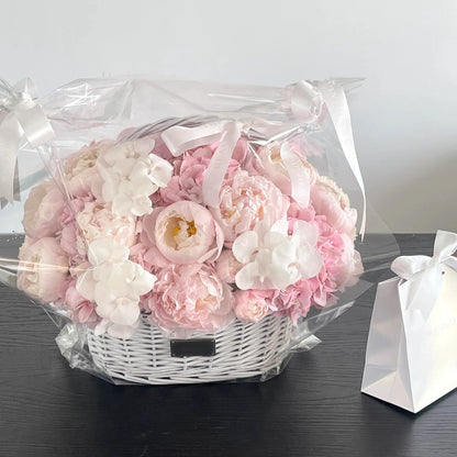 Pink and white preserved flower basket shown in clear wrapping beside a gift bag