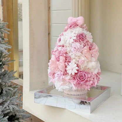 Pink and white preserved flower tree with roses on a reflective base by a window.