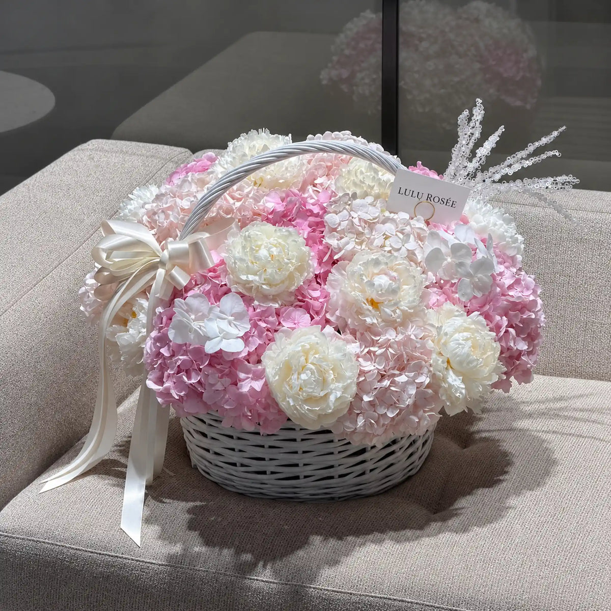 Pink preserved hydrangea and white peony basket displayed on a sofa with a cream bow