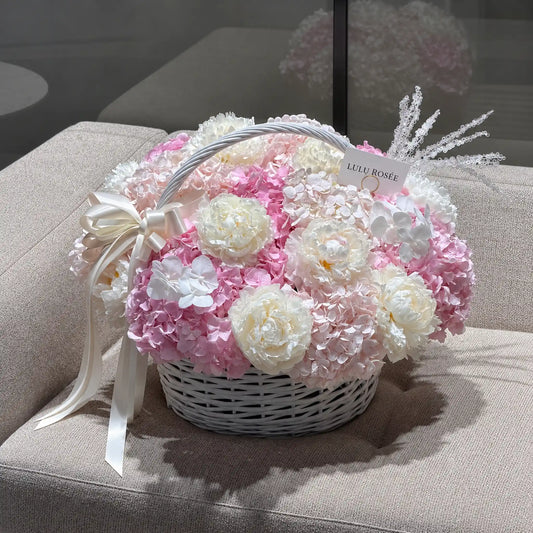 Pink preserved hydrangea and white peony basket displayed on a sofa with a cream bow