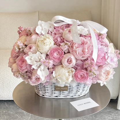 Pink preserved peonies and roses arranged in a white basket with a ribbon