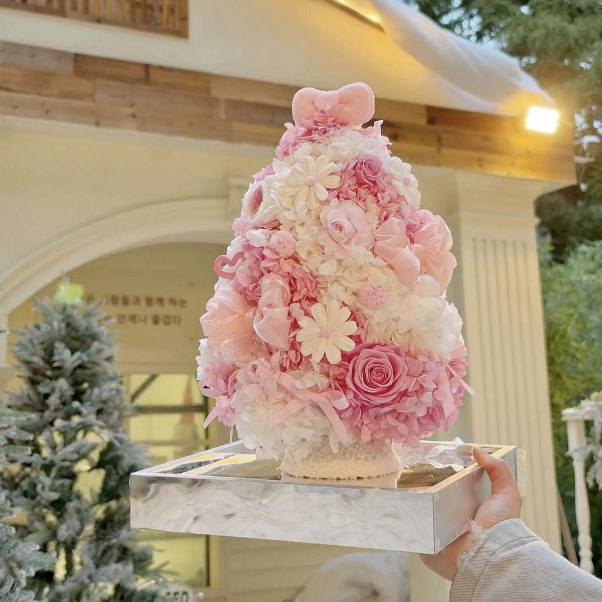 A person holds a pink preserved rose and hydrangea Christmas tree on a mirrored base.