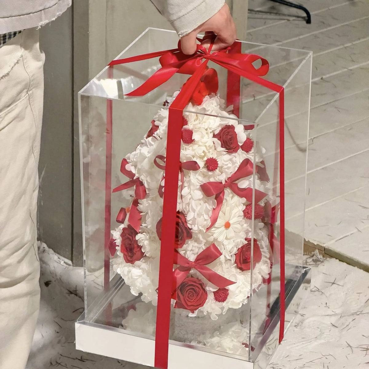 A person holding a clear gift box containing a red and white preserved flower tree.
