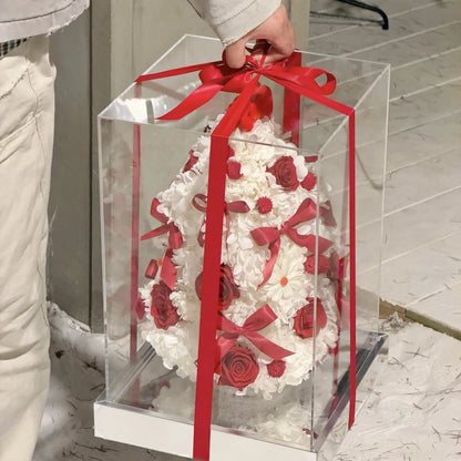 A person holding a clear gift box containing a red and white preserved flower tree.
