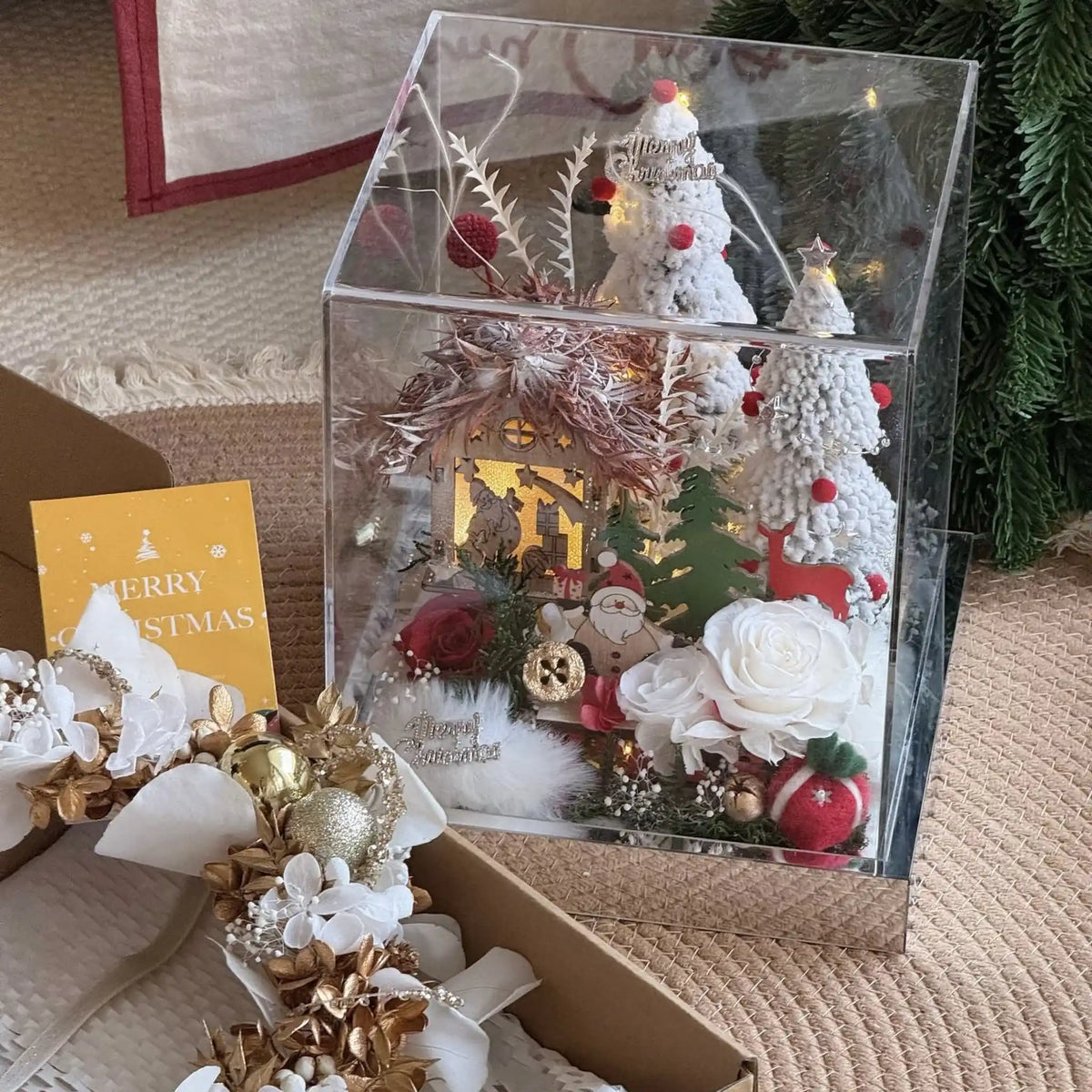 Red and white preserved roses frame a lit Christmas cottage inside a clear display box.