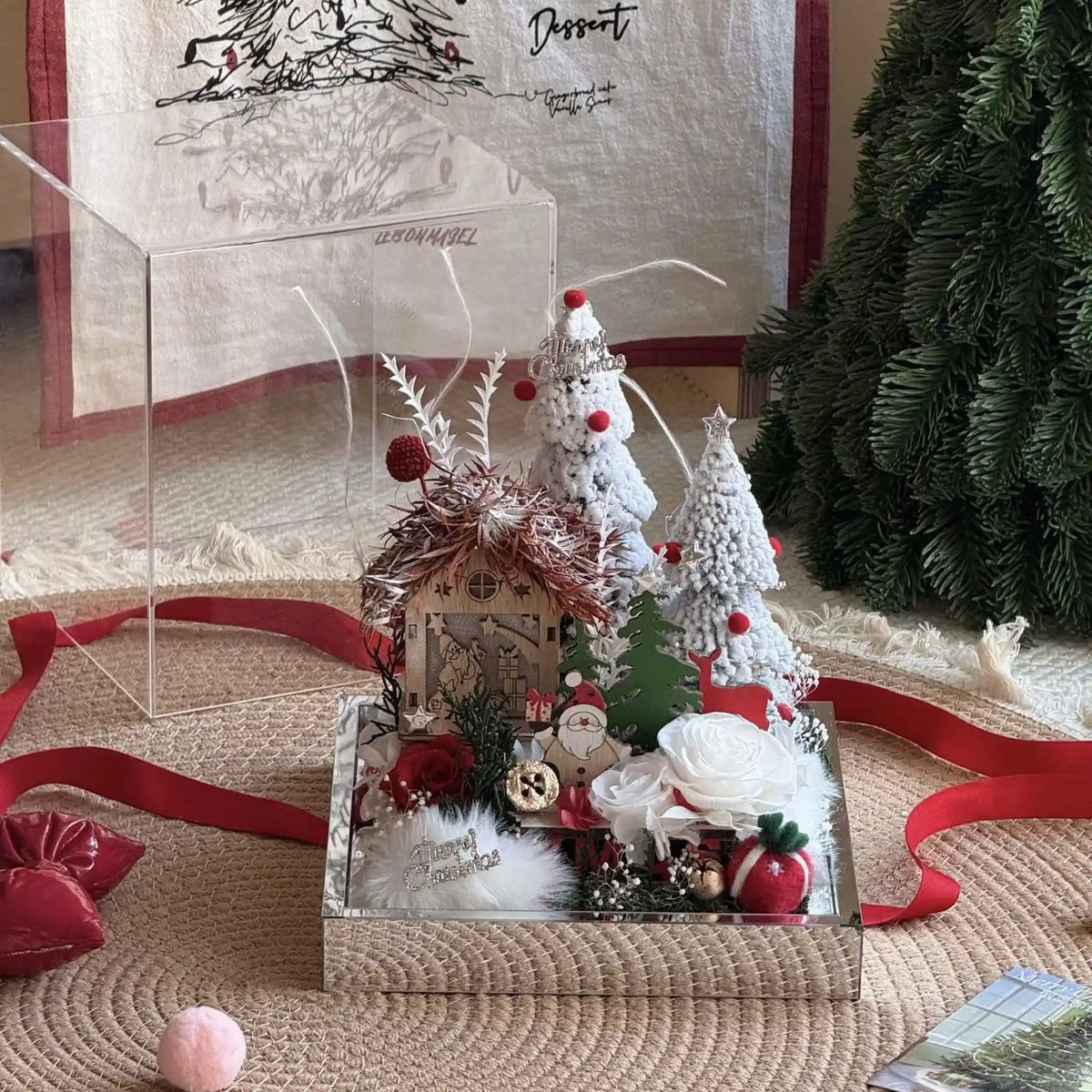 Preserved rose Christmas village displayed beside a red ribbon and evergreen tree.