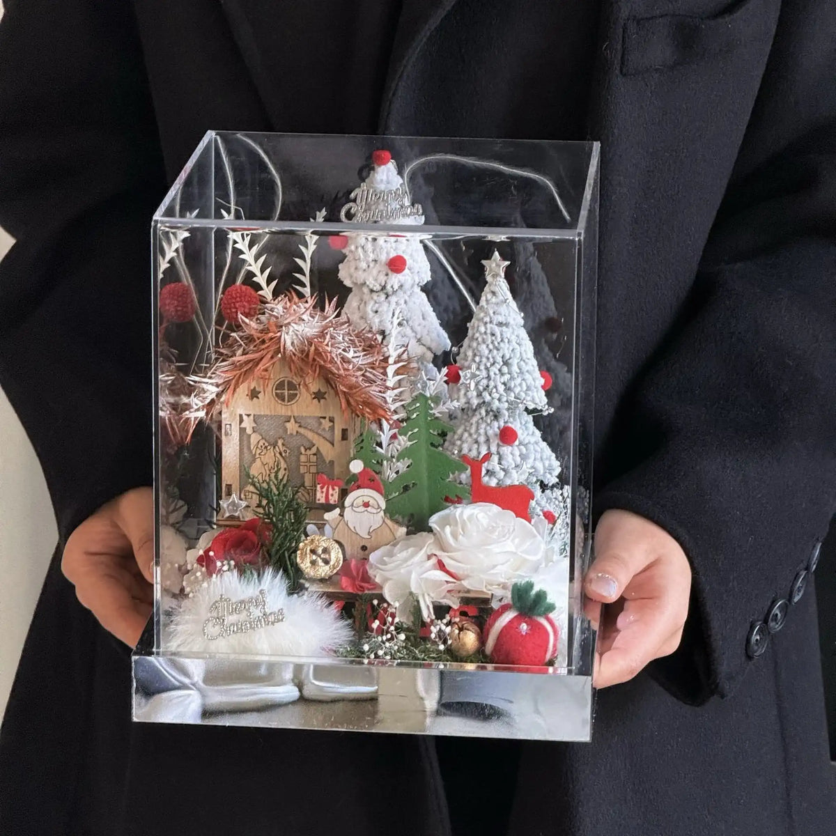 A person holds a clear display box containing a preserved rose Christmas village.