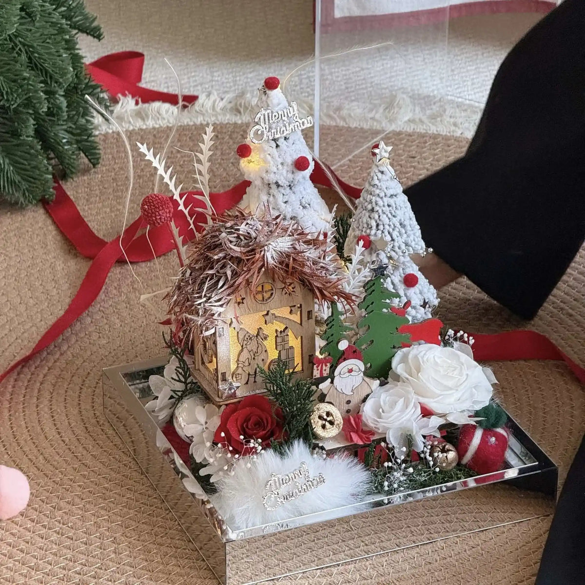 Open view of a Christmas village arrangement with preserved roses, snowy trees and a miniature cottage.